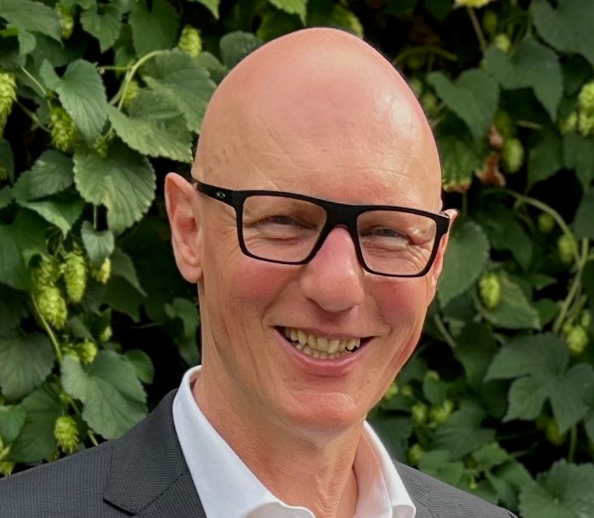 Smiling bald man wearing glasses and a suit in front of green foliage.