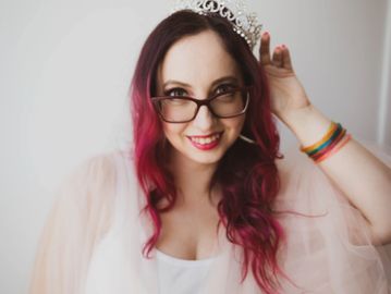 Young woman with pink hair wearing a tiara and glasses smiling.