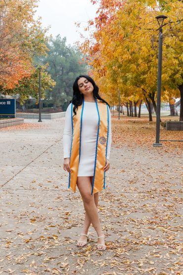 Woman in white dress and graduation stole standing on a leaf-covered path.