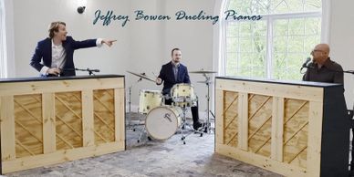 Three musicians performing with two pianos and a drum set in a bright room.