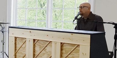 Man singing and playing piano near a large window.