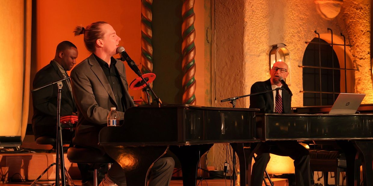 Two men in suits playing grand pianos and singing into microphones on a warmly lit stage.