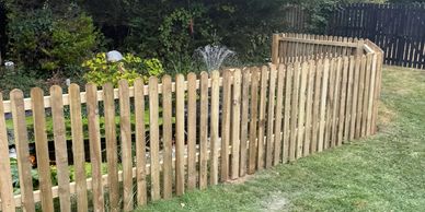 A wooden picket fence encloses a garden area with green plants and trees.