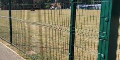 Green metal fence enclosing a grassy field under a blue sky.