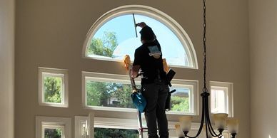 A worker on a ladder cleaning a high arched window inside a bright room.