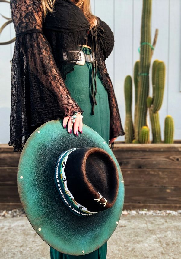 Woman holding a green and black wide-brimmed hat with a cactus background.