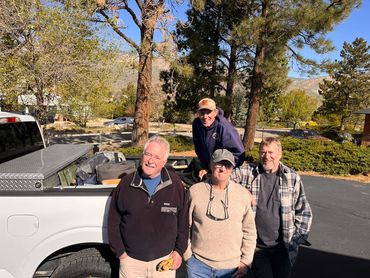 Four men posing outdoors near a pickup truck on a sunny day.