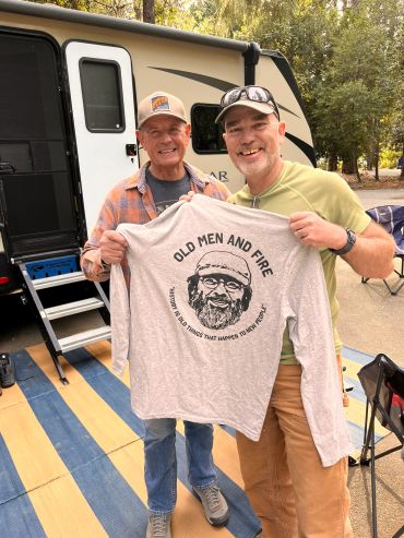 Two men smiling and holding a humorous shirt near a camper.