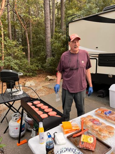 Man grilling burgers at a campsite with RV in background.