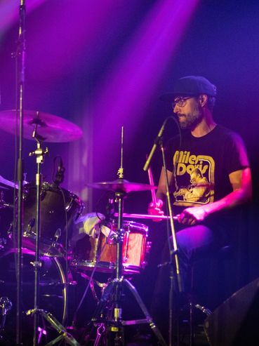 Drummer performing passionately on stage under purple lights.