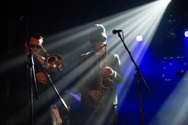 Two musicians playing saxophone and trombone under dramatic stage lights.