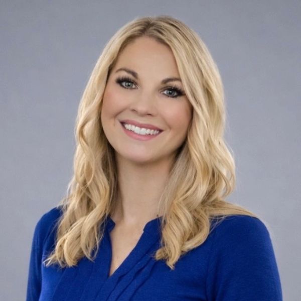 Smiling woman with blonde hair wearing a blue top against a gray background.