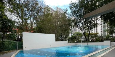 Outdoor swimming pool surrounded by trees and buildings under a bright sky.