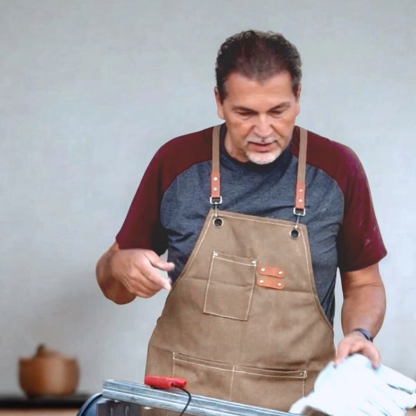 Man in apron holding gloves and inspecting metal pipes at a workbench.
