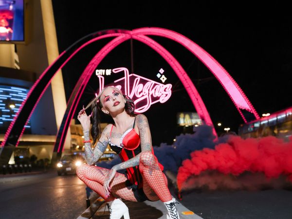 Tattooed woman in red fishnets and white boots poses under pink neon Las Vegas sign.