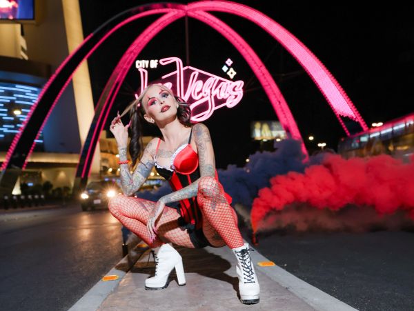 Tattooed woman in red fishnets and white boots poses under pink neon Las Vegas sign.