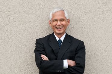 business headshot of a smiling man with white hair leaning against a stucco wall