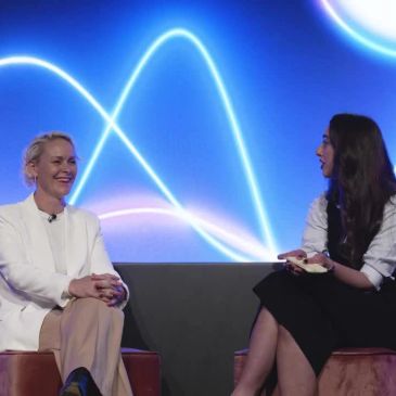 Two women engaged in a lively interview on a stage with a glowing blue background.