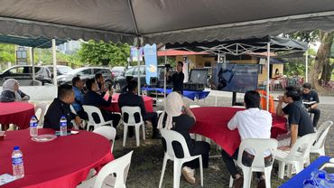 People attending an outdoor seminar under a tent with red and blue tables.