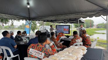 Firefighters attending an outdoor briefing under a tent near a water body.