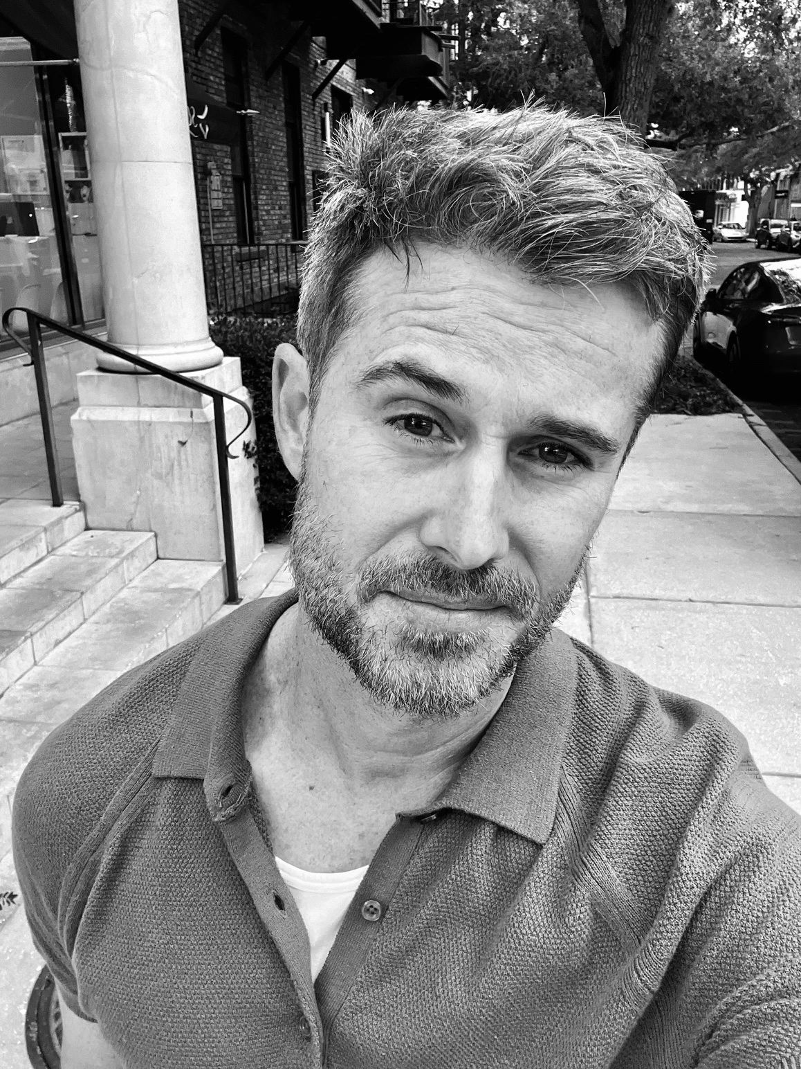 Black and white selfie of a man with a light beard on a city sidewalk.