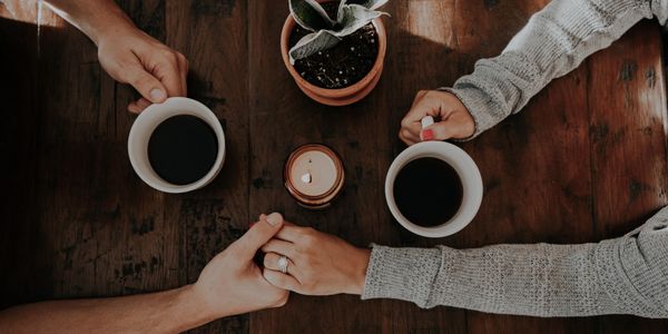 Two people sitting at a wood table drinking coffee out of white mugs while holding hands.