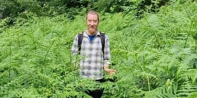 Man standing in a dense green fern forest wearing a checkered shirt and backpack.