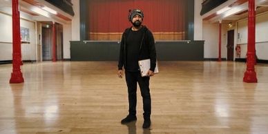 Man with headphones and laptop standing in an empty hall with wooden floor.
