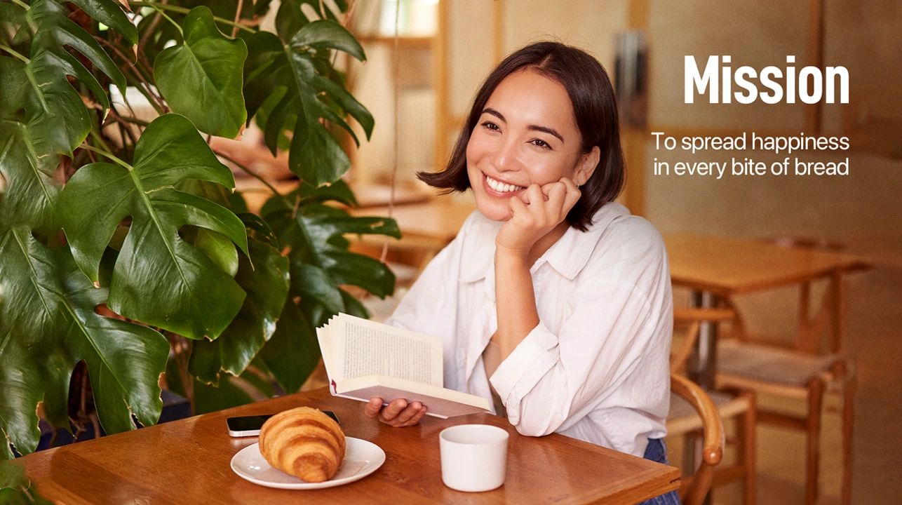 Smiling woman reading at a table with croissant and coffee, mission to spread happiness in bread.