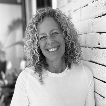 Smiling woman with curly hair leaning against a white brick wall.