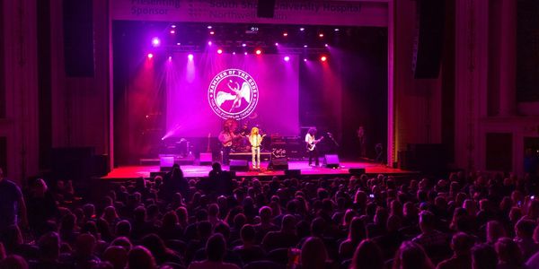 Band performing on stage with purple lighting and a large audience.