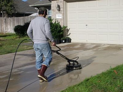 Man pressure washing a driveway wearing boots and a cap.