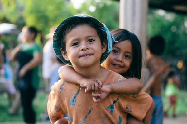 Two young children smiling, with one hugging the other from behind outdoors.