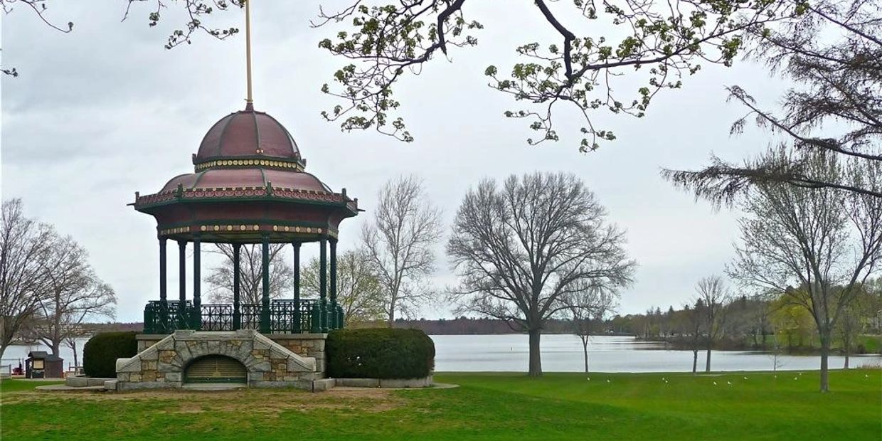 A decorative gazebo by a lake on a cloudy day with bare trees.