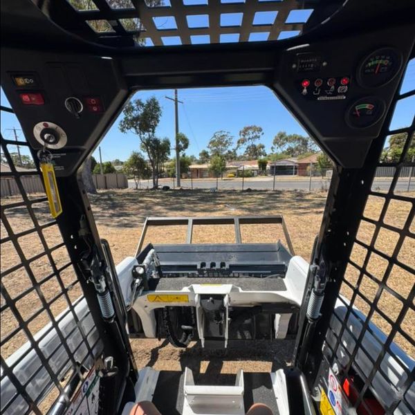View from inside a compact skid steer loader looking out onto a dry yard.