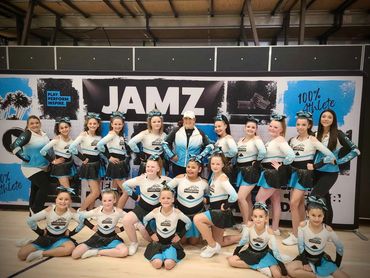 A cheerleading team in matching uniforms posing indoors in front of a JAMZ banner.
