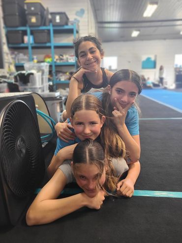 Four girls stacked playfully on a gym floor, smiling and posing cheerfully.