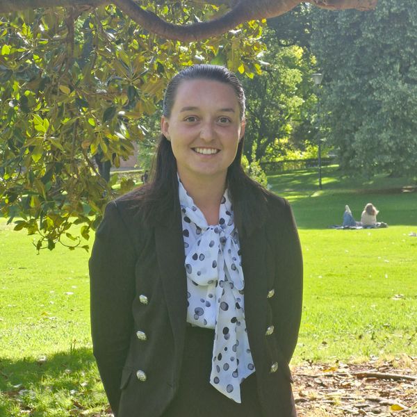 Smiling woman in a black blazer and polka dot blouse standing in a sunny park.