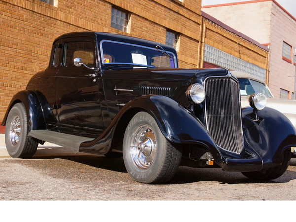 A shiny black vintage car at a past Munising Bay Cruisers car show with a brick building backdrop.