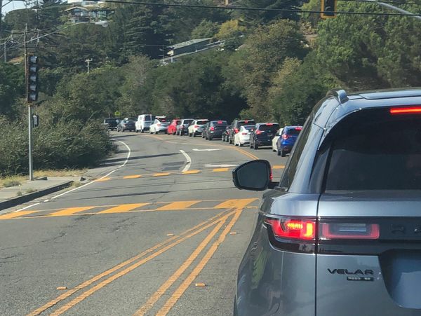 Cars lined up on a hilly road with houses in the background.