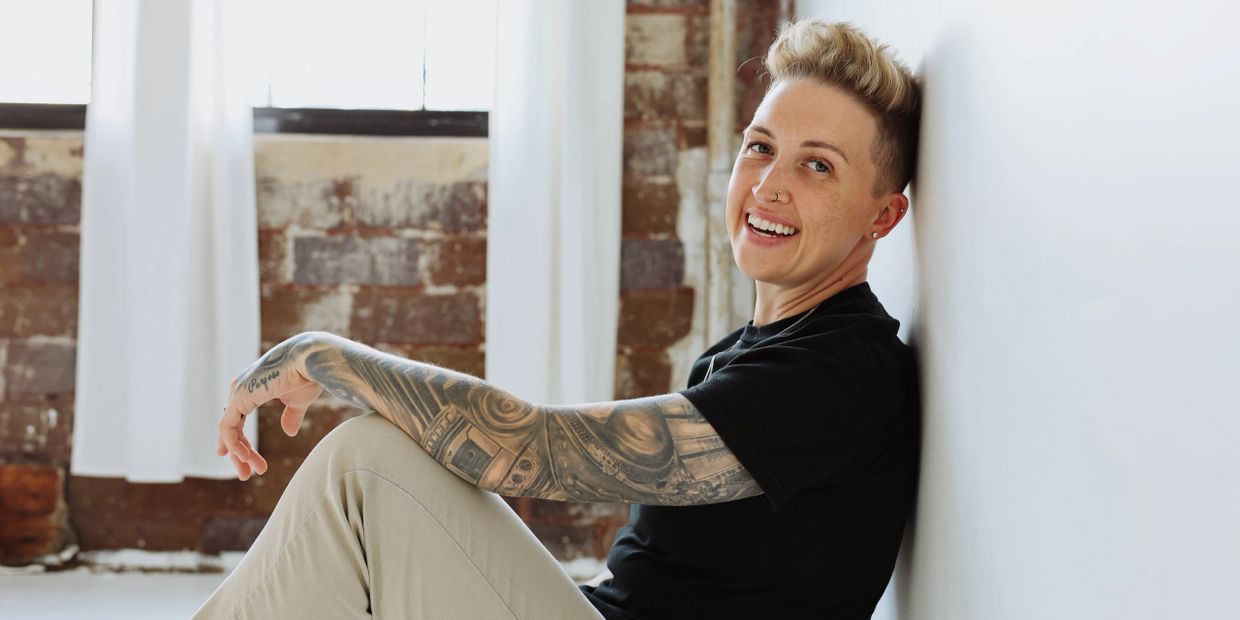 Smiling person with tattoos sits relaxed against a white wall in a bright room.