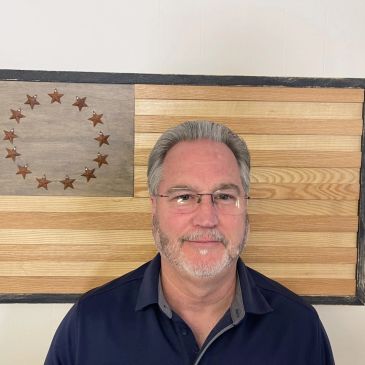Man with gray hair and glasses in front of wooden American flag.