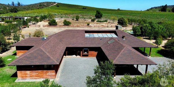 A large wooden house with a brown roof surrounded by greenery and hills.