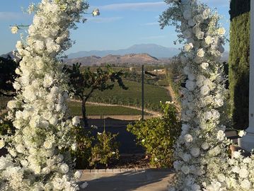 White rose and baby’s breath silk floral archway with vineyard and mountains background at Mount Palomar winery wedding venue and n Temecula CA.