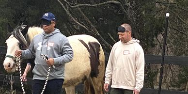 Two men walking a horse in an outdoor setting, one man wearing an eye patch.
