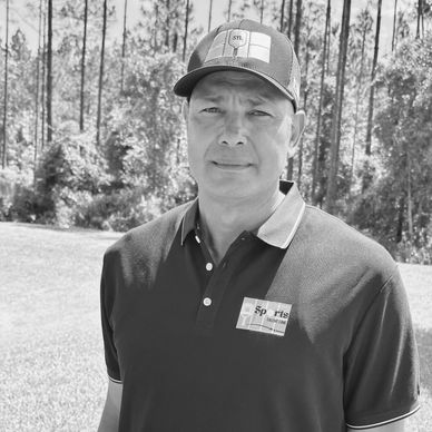 Man wearing a cap and polo shirt standing outdoors in a wooded area.