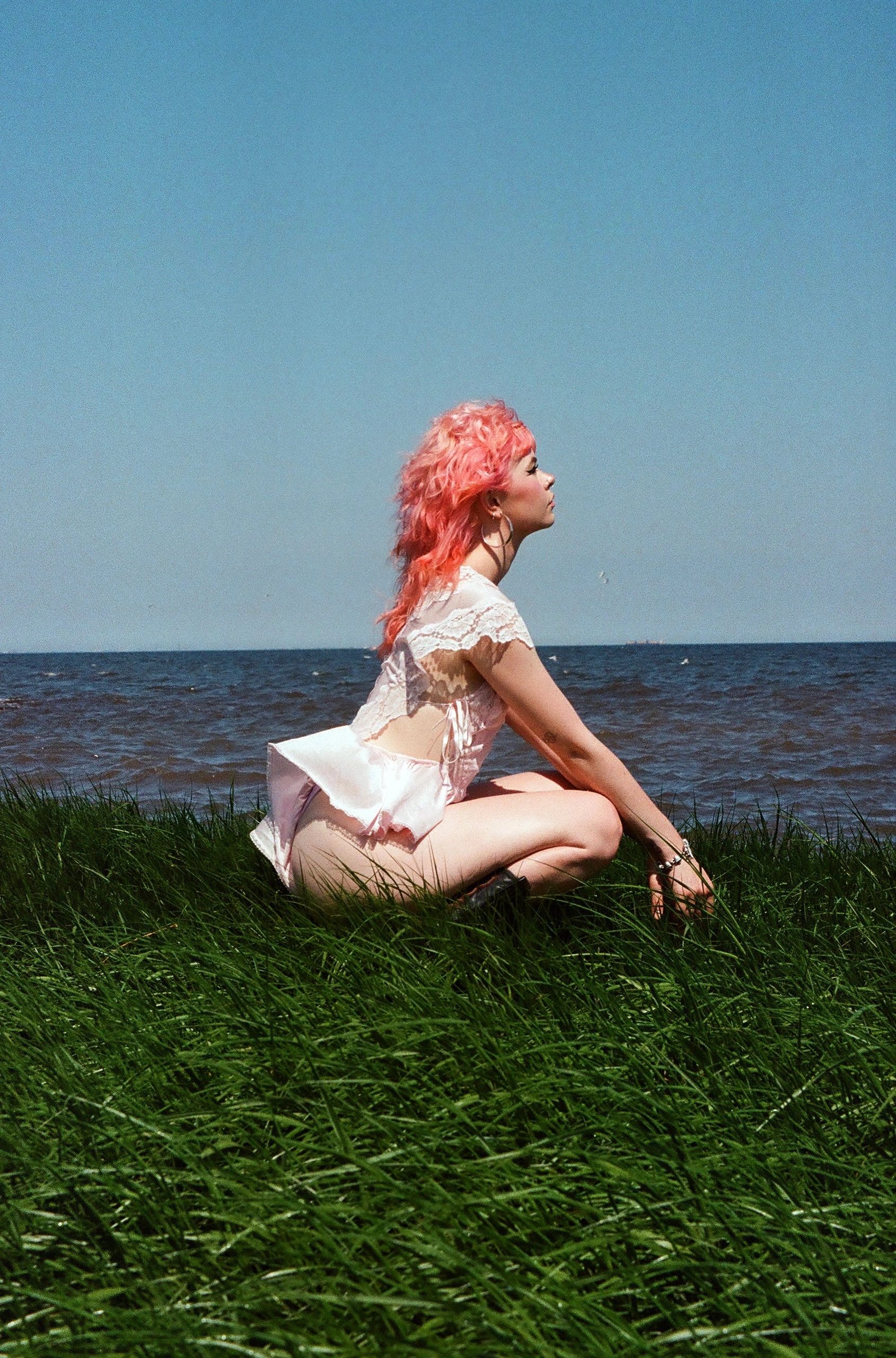Person with pink hair sitting by the lake in grass.
