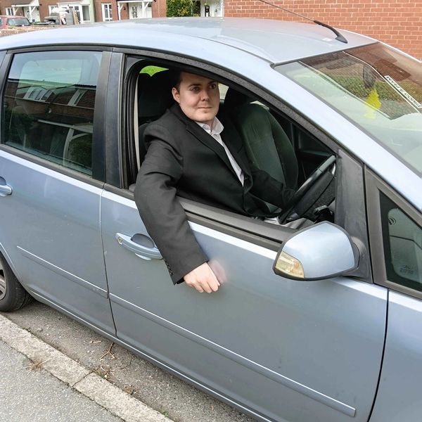 Person in a black suit sitting in a parked silver car with the window down.