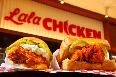 Two crispy fried chicken sandwiches with pickles and sauce in front of a Lala Chicken sign.