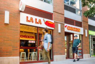 People walking past La La Chicken restaurant with a bright sign.